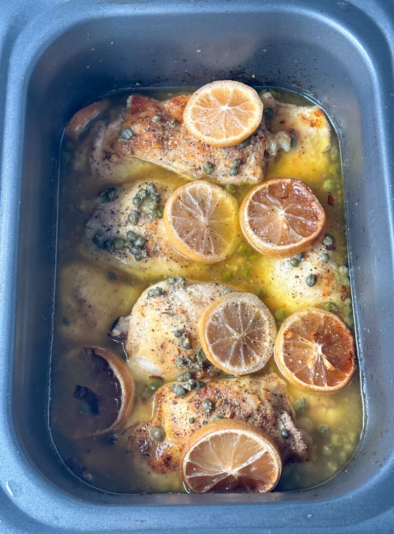 slow cooked chicken piccata in a gray rectangular crockpot topped with lemon rounds and capers