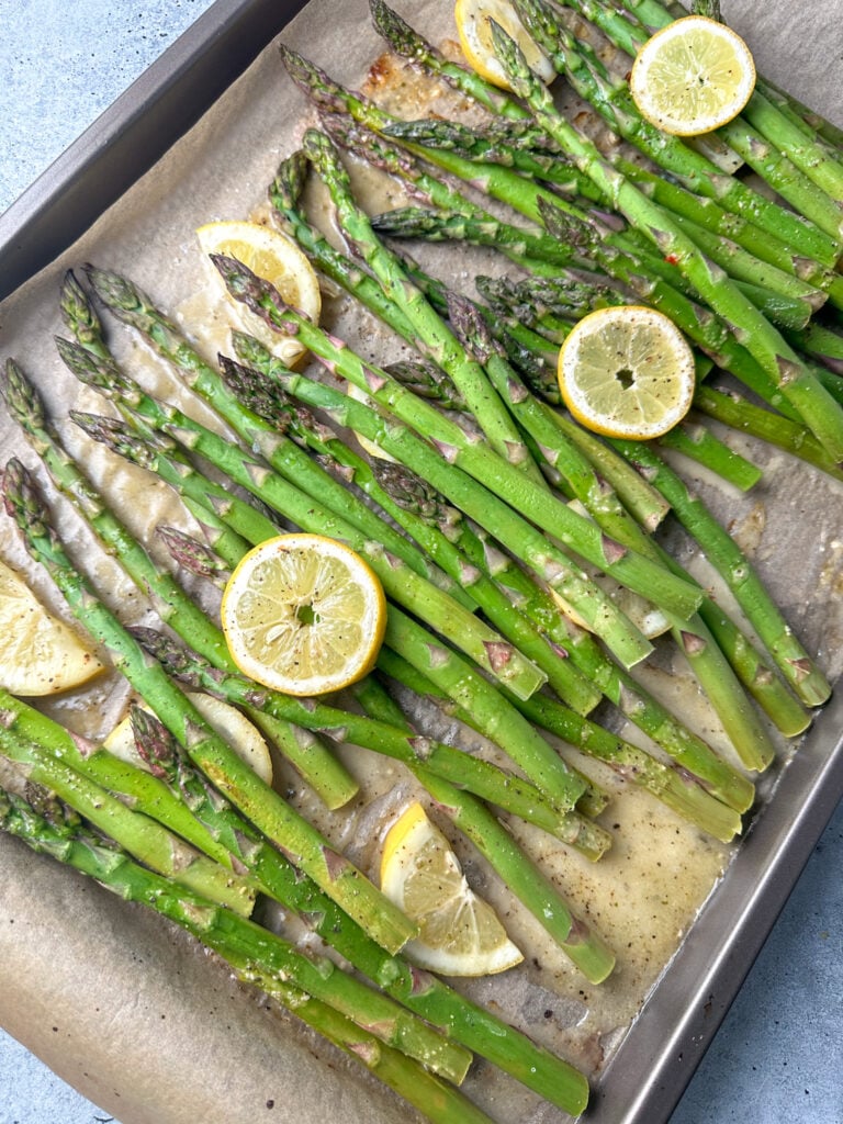 asparagus on a baking sheet with brown parchment with Italian dressing and lemon rounds