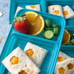 a blue lunchbox with a cheese quesadilla that has flower cut-outs on it served with fresh fruit