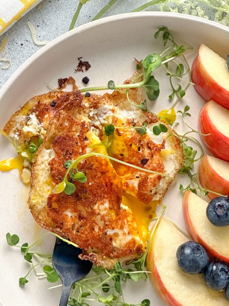 crispy cheese fried egg on a white plate with a slightly runny egg yolk and fresh sprouts served with apples and blueberries