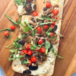 Rectangular shaped flatbread pizza topped with Boursin cheese, caramelized onion, mushroom, and fresh arugula on a wooden pizza tray.