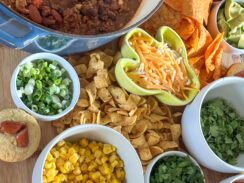 chili board featuring cheesy chili, heart shaped cocktail sausage cornbread muffins, corn, cilantro, jalapeño, diced red peppers, Fritos, Doritos, shredded cheese, avocado, black olives on a large wooden serving tray with a wooden spoon in the chili ready for game day