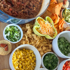 chili board featuring cheesy chili, heart shaped cocktail sausage cornbread muffins, corn, cilantro, jalapeño, diced red peppers, Fritos, Doritos, shredded cheese, avocado, black olives on a large wooden serving tray with a wooden spoon in the chili ready for game day