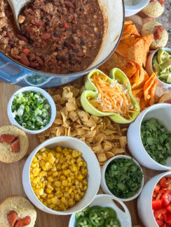 chili board featuring cheesy chili, heart shaped cocktail sausage cornbread muffins, corn, cilantro, jalapeño, diced red peppers, Fritos, Doritos, shredded cheese, avocado, black olives on a large wooden serving tray with a wooden spoon in the chili ready for game day