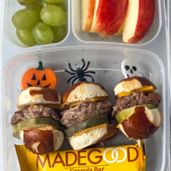 soft pretzel bites with a burger, pickle and cheese held together by halloween toothpicks in a lunchbox with apple slices, green grapes, and granola bar
