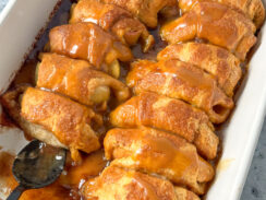 crescent rolls with apples inside topped with a buttery cinnamon glaze with caramel dip drizzled over in a white baking dish.