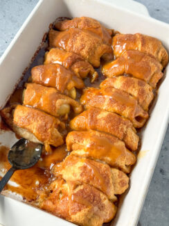 crescent rolls with apples inside topped with a buttery cinnamon glaze with caramel dip drizzled over in a white baking dish.