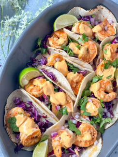 shrimp in toasted corn tortillas served with purple cabbage, cilantro leaves, lime wedges, and a creamy buffalo ranch sauce in a gray serving platter.