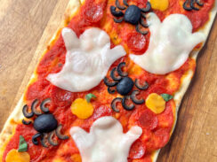 rectangle flatbread pizza on a wooden pizza serving tray topped with pizza sauce, mini pepperonis, melted mozzarella cheese ghost cut-outs, cheddar cheese pumpkin cut-outs, and spiders made from black olives.