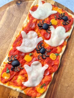 rectangle flatbread pizza on a wooden pizza serving tray topped with pizza sauce, mini pepperonis, melted mozzarella cheese ghost cut-outs, cheddar cheese pumpkin cut-outs, and spiders made from black olives.