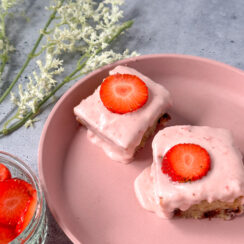 two banana cake brownies on a pink plate with strawberry frosting and a fresh slice of strawberry on top