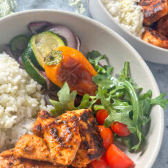 bite sized blackened salmon in a white bowl with white rice, roasted vegetables, cherry tomatoes, and white rice