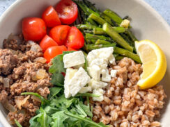 farro, roasted asparagus, cherry tomatoes, ground beef, arugula, feta cheese, and lime wedge in a white bowl