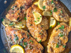 flour dredged crispy chicken breasts in a large skillet with a brown butter sauce topped with capers, lemon wedges, and parsley leaves.
