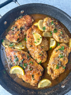 flour dredged crispy chicken breasts in a large skillet with a brown butter sauce topped with capers, lemon wedges, and parsley leaves.