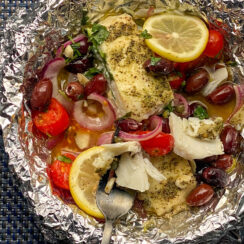 white fish in a bowl with foil topped with lemon slices, cherry tomatoes, red onion, Kalamata olives, parsely, and seasoning.