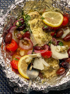 white fish in a bowl with foil topped with lemon slices, cherry tomatoes, red onion, Kalamata olives, parsely, and seasoning.