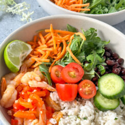 shrimp, coconut cilantro rice, cucumber, black beans, arugula, shredded carrots, and lime wedge in a white serving bowl