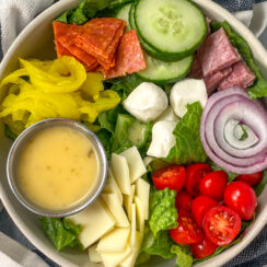 chopped romaine lettuce, pepperoni, salami, cucumber, red onion, cherry tomatoes, banana peppers, and mozzarella balls in a white bowl served with Italian dressing