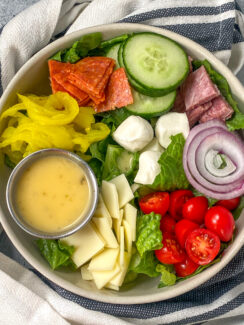 chopped romaine lettuce, pepperoni, salami, cucumber, red onion, cherry tomatoes, banana peppers, and mozzarella balls in a white bowl served with Italian dressing