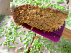 A slice of banana bread with vanilla frosting and green sprinkles on purple spatula in a white baking dish