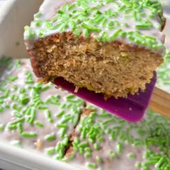 A slice of banana bread with vanilla frosting and green sprinkles on purple spatula in a white baking dish