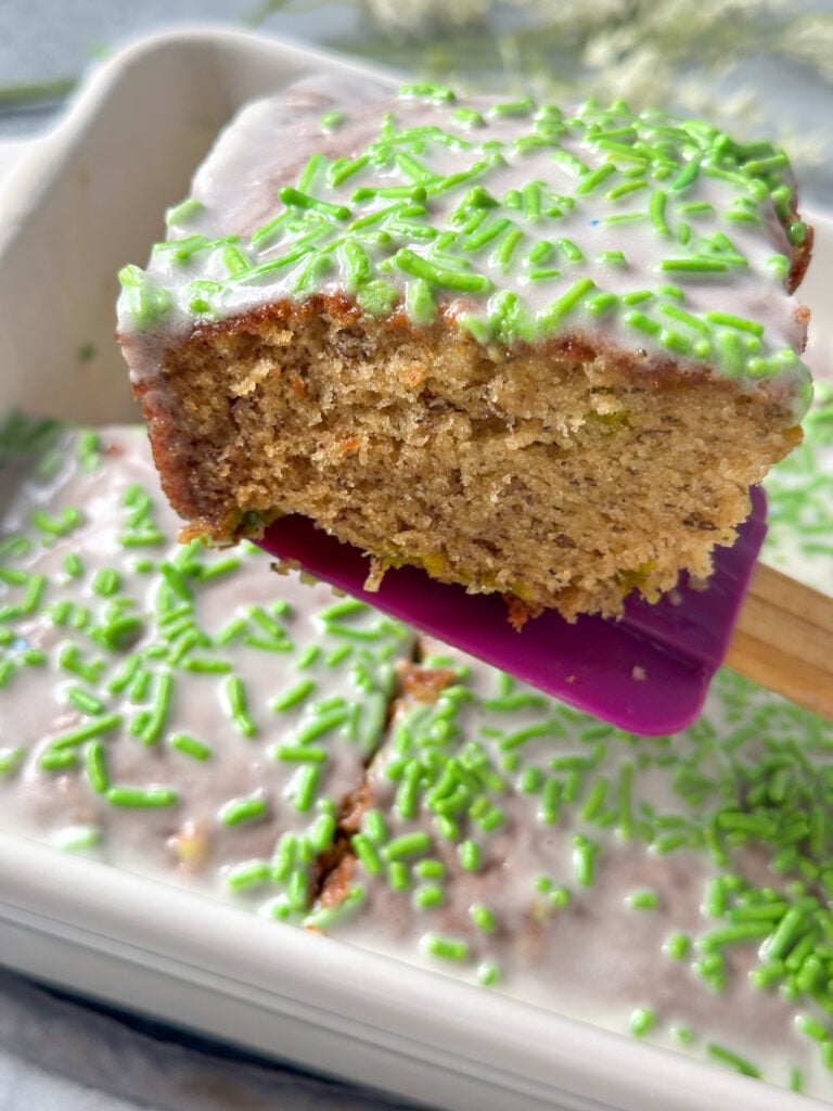 A slice of banana bread with vanilla frosting and green sprinkles on purple spatula in a white baking dish