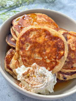 golden cottage cheese and brown sugar pancakes in a white bowl served with whipped cream and a sprinkle of cinnamon with maple syrup drizzle.