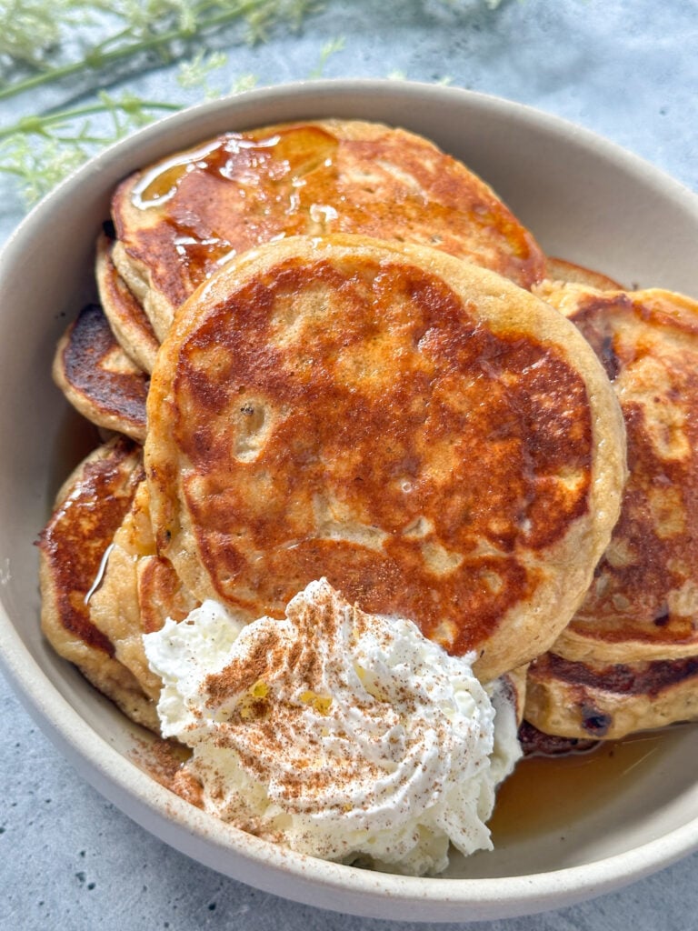 golden cottage cheese and brown sugar pancakes in a white bowl served with whipped cream and a sprinkle of cinnamon with maple syrup drizzle.