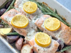 4 salmon fillets in a white baking dish on top of baby potatoes and green beans with a creamy dill sauce and lemon rounds on top
