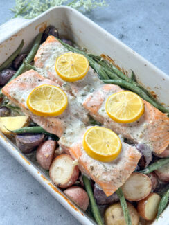 4 salmon fillets in a white baking dish on top of baby potatoes and green beans with a creamy dill sauce and lemon rounds on top