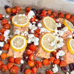 salmon fillets on a sheet pan with roasted cherry tomatoes and Kalamata olives and topped with lemon wedges and crumbled feta cheese