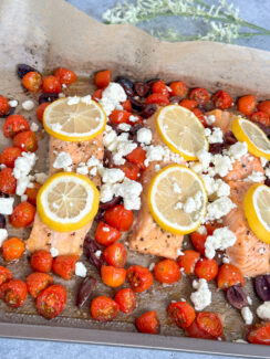 salmon fillets on a sheet pan with roasted cherry tomatoes and Kalamata olives and topped with lemon wedges and crumbled feta cheese
