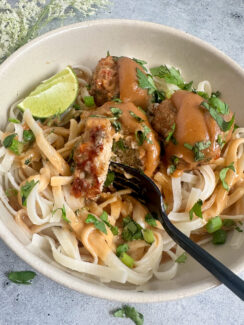 thai chicken meatballs with a creamy peanut sauce drizzle over served with thai noodles, fresh cilantro and lime in a white bowl