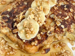 banana crunch french toast on a white plate with fresh banana slices, granola, whipped cream, and maple syrup.