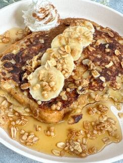 banana crunch french toast on a white plate with fresh banana slices, granola, whipped cream, and maple syrup.