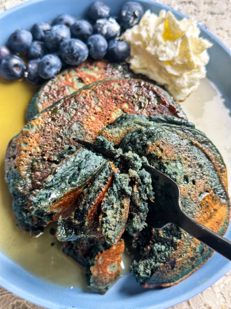 blender blueberry oatmeal pancakes with a fork cut into them showing their naturally blue color on a blue plate with maple syrup drizzled on top along with fresh blueberries and coconut milk whipped cream