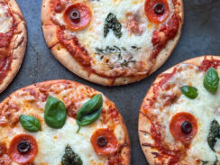 day of the dead skull pizzas made with mini boboli crusts, pizza sauce, provolone cheese, shredded mozzarella cheese, basil leaves, pepperoni, black olives, and thyme leaves on a gray baking dish