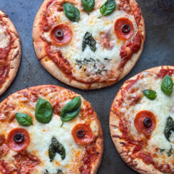 day of the dead skull pizzas made with mini boboli crusts, pizza sauce, provolone cheese, shredded mozzarella cheese, basil leaves, pepperoni, black olives, and thyme leaves on a gray baking dish