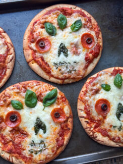 day of the dead skull pizzas made with mini boboli crusts, pizza sauce, provolone cheese, shredded mozzarella cheese, basil leaves, pepperoni, black olives, and thyme leaves on a gray baking dish