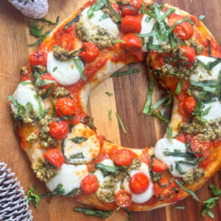 holiday pizza wreath made with refrigerated pizza dough, fresh mozzarella balls, cherry tomatoes, pizza sauce, and fresh shredded basil leaves on a wooden pizza board with winter pinecones