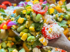 chopped salad with edamame, corn, avocado, cherry tomatoes, red onion, and crumbled feta on a wooden spoon with hot honey vinaigrette on top