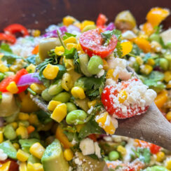 chopped salad with edamame, corn, avocado, cherry tomatoes, red onion, and crumbled feta on a wooden spoon with hot honey vinaigrette on top