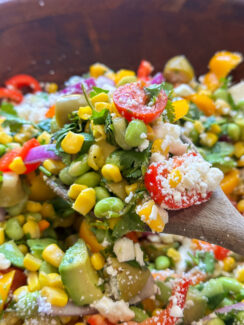 chopped salad with edamame, corn, avocado, cherry tomatoes, red onion, and crumbled feta on a wooden spoon with hot honey vinaigrette on top