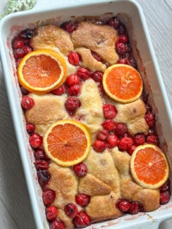 cranberry orange cream dumplings made with crescent rolls in a white baking dish with a brown sugar glaze, baked cranberries, and orange slices