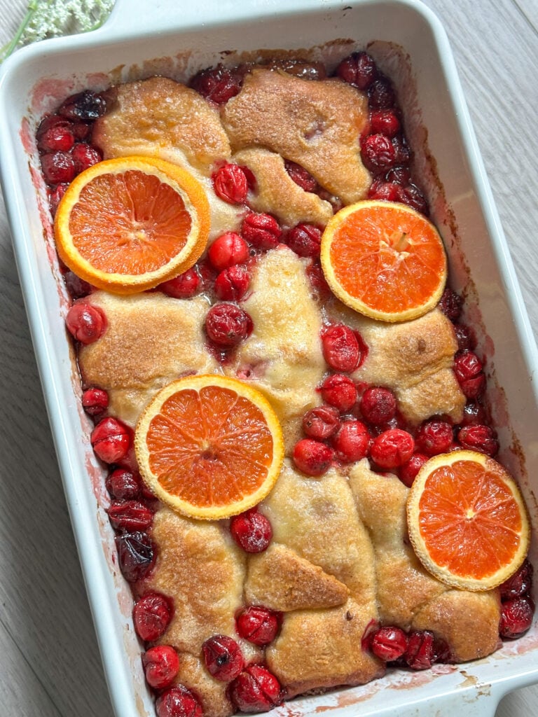 cranberry orange cream dumplings made with crescent rolls in a white baking dish with a brown sugar glaze, baked cranberries, and orange slices
