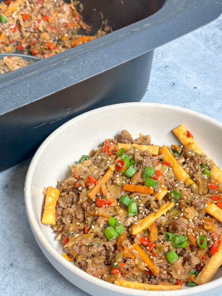 crockpot egg roll in a bowl served in a white serving bowl next to a square, gray slow cooker topped with crunchy Chinese noodles and fresh green onion.